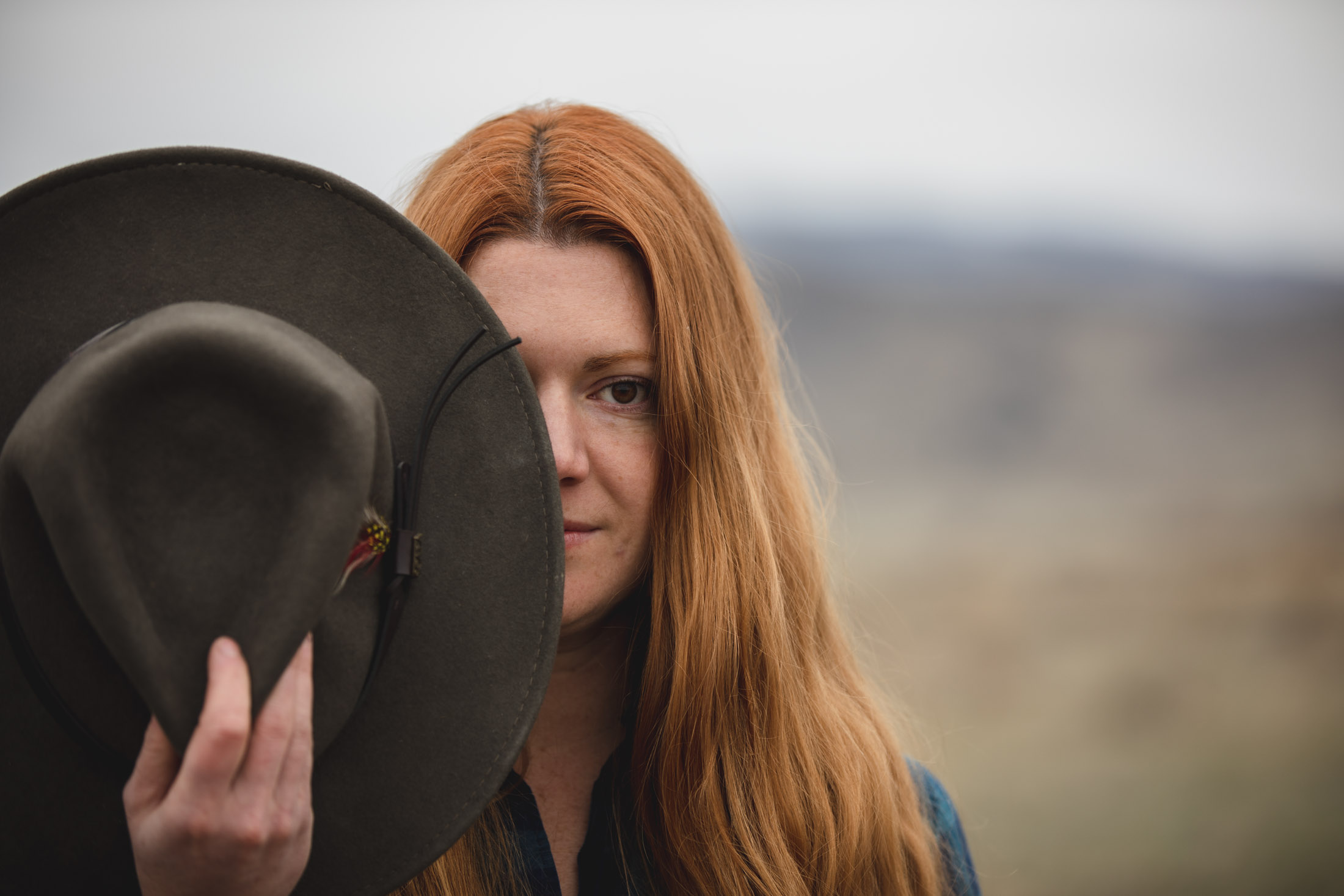 Outdoor portrait of a red haired woman covering up one eye with her hat - John Robson Photography