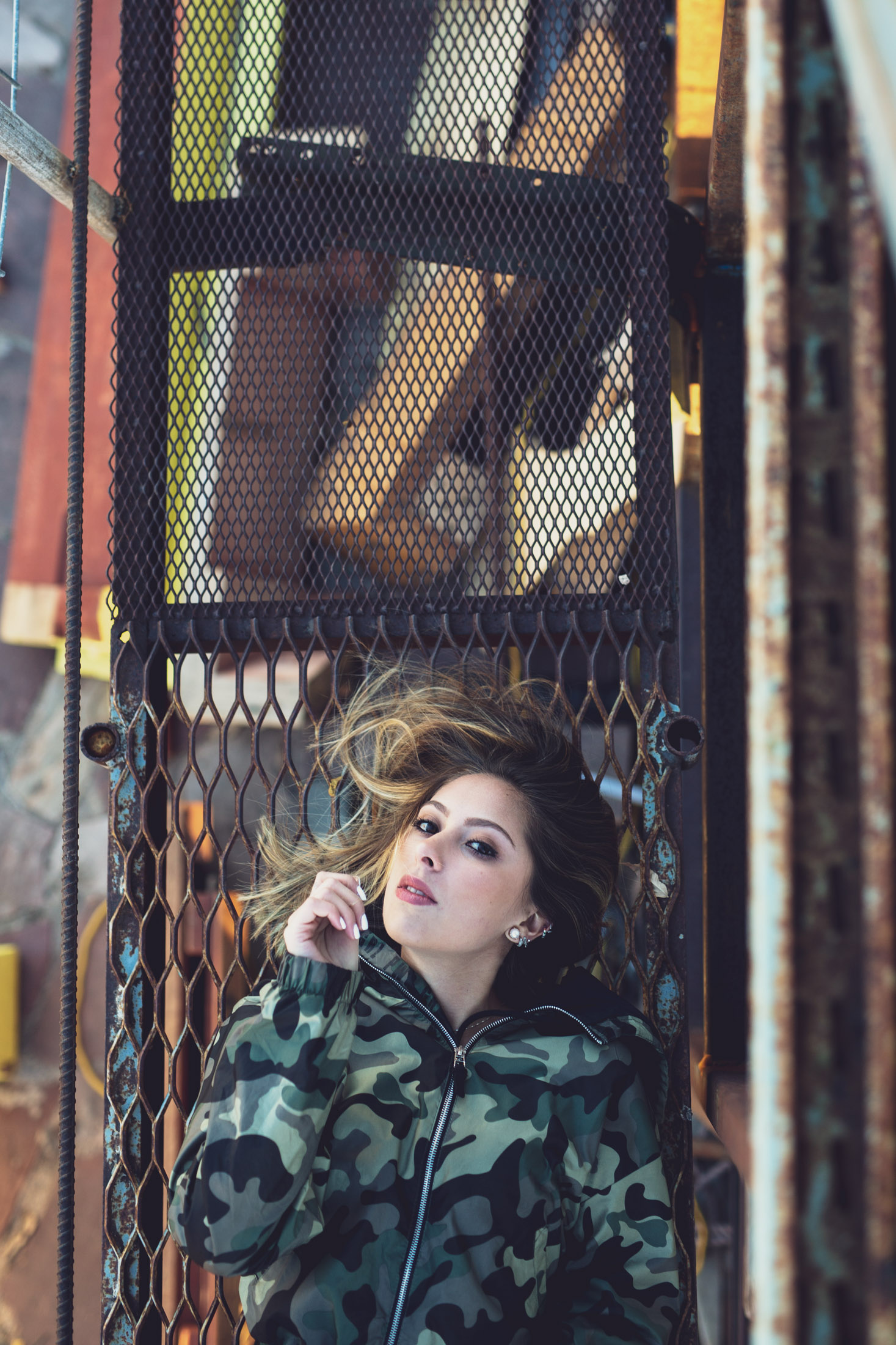 Portrait of a young woman wearing a camo jacket laying on a metal grate - John Robson Photography