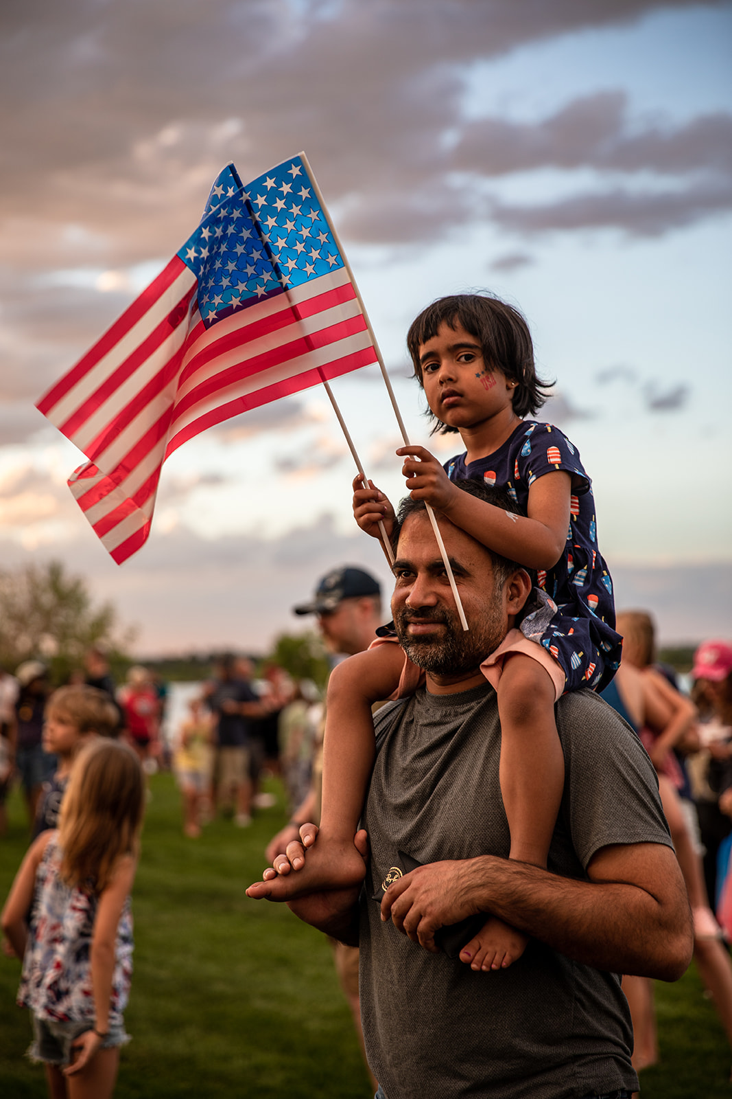 Girl sitting on her father shoulders and holding two American flags at a July 4th event- John Robson Photography
